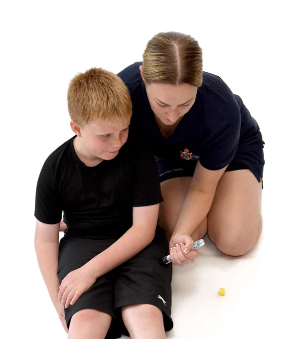 A person is kneeling on the floor administering an injection into the leg of a seated child. The child is wearing a black shirt and shorts. The background is white.