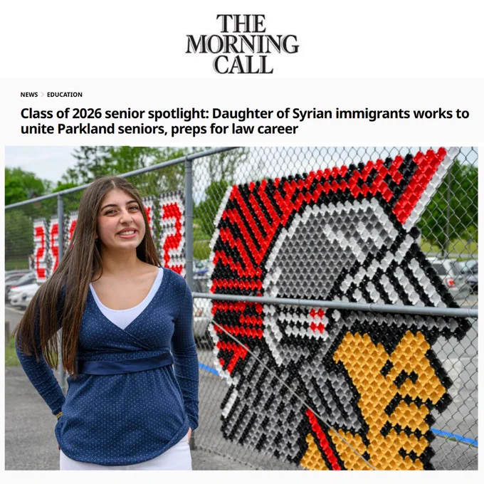 A smiling woman with long hair stands next to a fence with a mosaic image of a helmet.