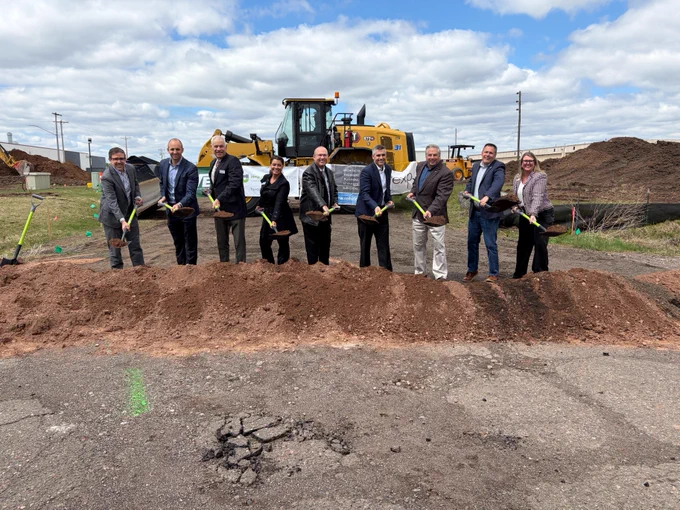 A group of individuals stand in a line holding shovels at a groundbreaking ceremony. They are in front of a pile of dirt with construction equipment in the background. The sky is partly cloudy.
