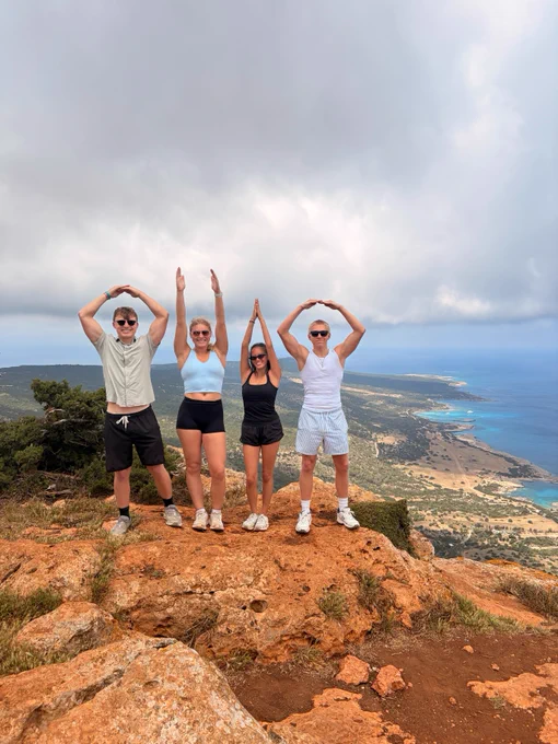 Four people stand on a rocky cliff with their arms forming the letters "O", "H", "I", and "O". The background features a scenic view of a coastline with the sea, hills, and cloudy sky.