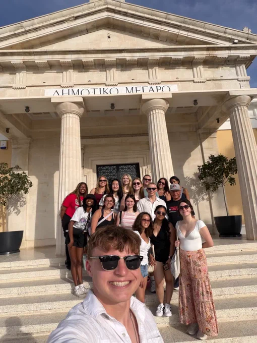 A group of people are gathered on the steps of a building labeled "ΔΗΜΟΤΙΚΟ ΜΕΓΑΡΟ," which translates to "City Hall" in English. The building features classical architectural elements such as columns. The group is taking a photo in bright sunlight.