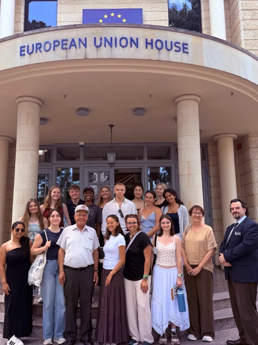 A group of people stand in front of the European Union House. They are posing on stairs beneath a sign that reads "European Union House." The group appears diverse and is smiling.