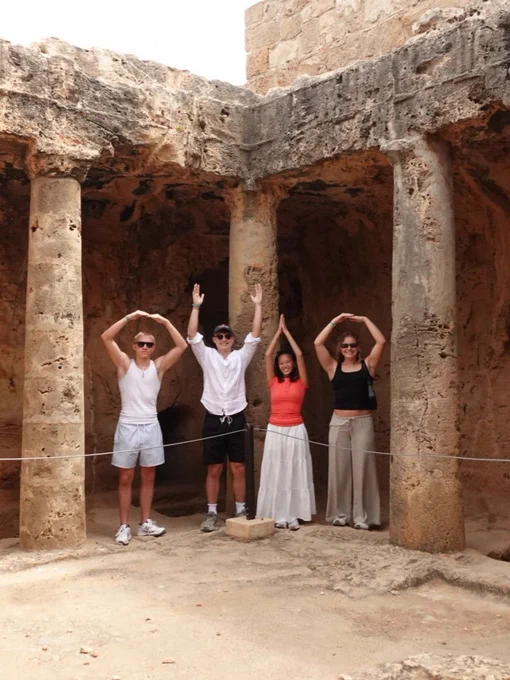 Four people stand between ancient stone columns, forming the word "OHIO" with their arms. They are inside a historic ruin site.