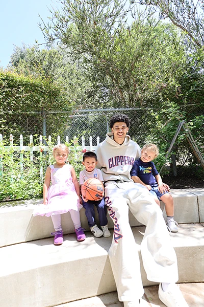 Kobe Sanders wearing a Los Angeles Clippers hoodie is sitting on outdoor steps with three young children. One child holds a basketball. The background features a fence and greenery.