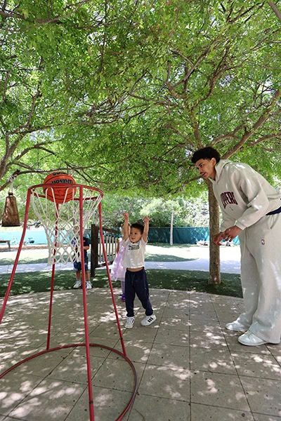 A child is shooting a basketball into a hoop while an adult in a sports tracksuit observes. They are outdoors on a tiled patio, surrounded by green leafy trees.