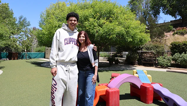 An individual in a Clippers tracksuit stands beside another person on a playground with colorful play equipment. Green trees and a grassy area are in the background.