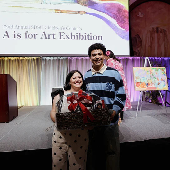 Two people stand smiling on stage at the “A is for Art Exhibition.” One holds a gift basket with a large red bow. In the background, there's an abstract painting on an easel and a colorful backdrop on stage.