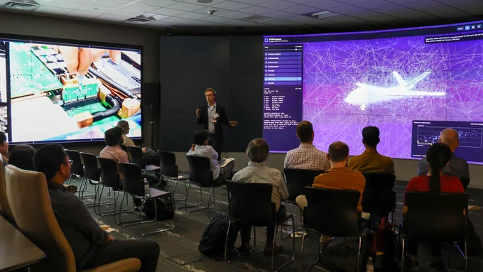 A presenter stands between two large display walls. One screen shows computer hardware imagery and the other displays a complex aircraft simulation rendered with interconnected computational data.