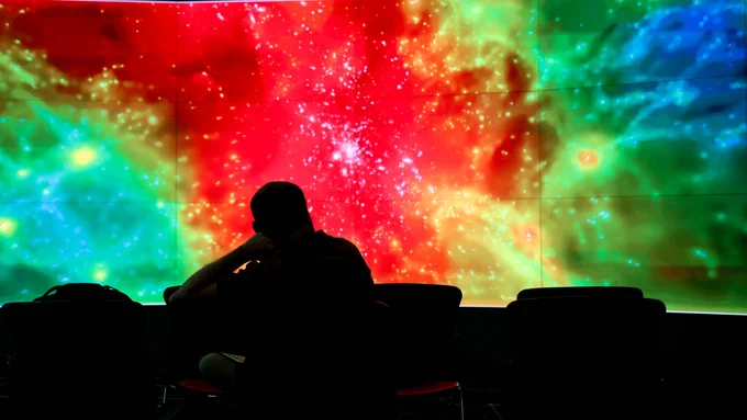 A silhouetted person sits facing a wall-sized scientific visualization depicting a colorful cosmic structure with bright red, green and blue regions. The immersive display is part of the data visualization showcase at the 2026 Oak Ridge Leadership Computing Facility User Summit.