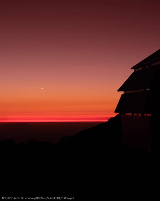 Thin crescent moon above a vivid red‑orange twilight horizon. Dark silhouette of an observatory at right