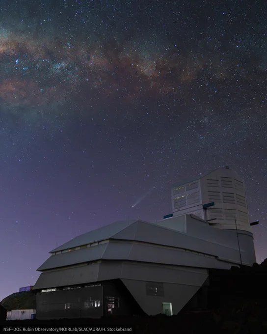 Purple-hued night sky over an angular white observatory. The Milky Way stretches overhead and a small comet with a tail sits above the observatory.