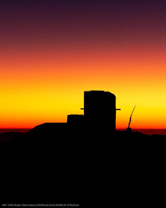 Silhouette of an angular observatory building against a vibrant yellow sunset sky fading to purple. A crane with arm extended is silhouetted to the right.
