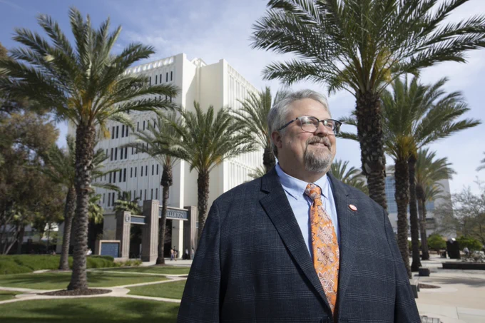 Person wearing a suit and patterned tie stands outside Cal State Fullerton, surrounded by palm trees and modern buildings. They appear content and are looking into the distance.