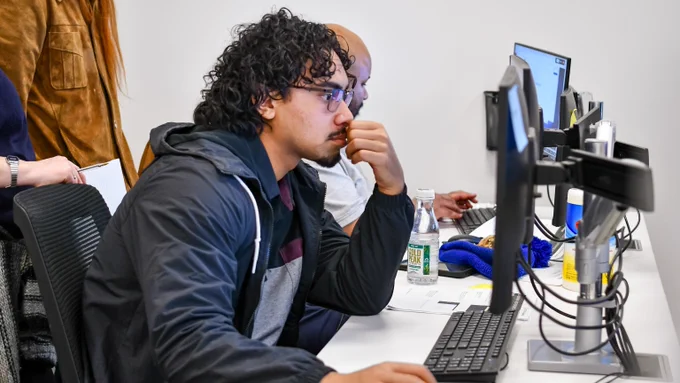 Man with curly hair and glasses intently working on a desktop computer in an office setting.
