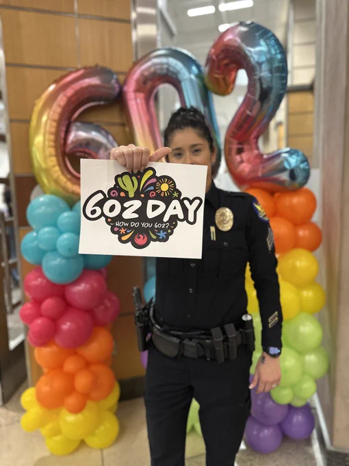 A person in a police uniform is holding a sign that reads "602 Day, How Do You 602?" in front of colorful balloons shaped as "602.
