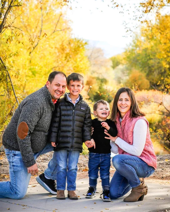 This image displays a group of four people outdoors in a park, surrounded by colorful autumn trees. All are smiling and appear to be enjoying a pleasant day.