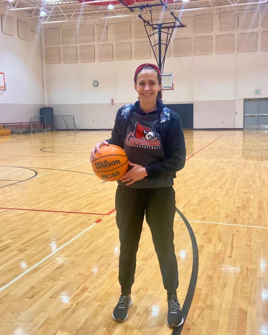 A person stands on an indoor basketball court holding a Wilson basketball. The person is wearing a Cardinals basketball sweatshirt and dark pants. Basketball hoops are visible in the background.