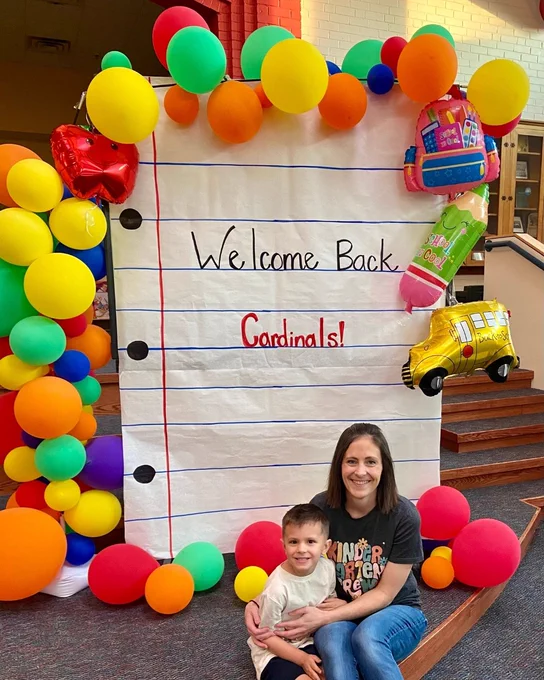 A person and a child sit in front of a large, decorated backdrop resembling a sheet of lined paper. The backdrop says "Welcome Back Cardinals!" It is adorned with colorful balloons, including a school bus balloon. Bright, festive atmosphere suggests a school setting.