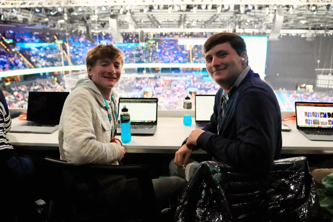 Two people sitting at a desk in a busy indoor arena with laptops and blue energy drinks.