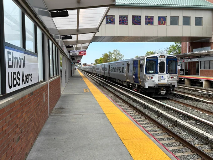 An LIRR M9 train running along the middle track past Elmont-UBS Arena station during the daytime.