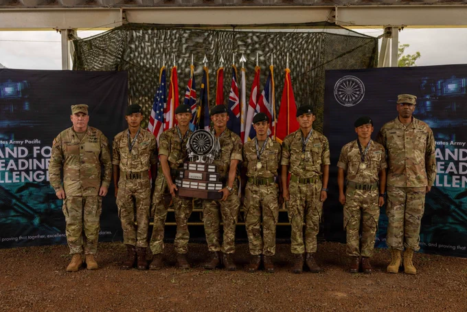 A group of uniformed military personnel stands in a line outdoors under a shelter. They are holding a wooden and metal trophy. Behind them are flags, including those of the United States and the United Kingdom. Two large banners are displayed on either side. The ground is covered with brown soil.