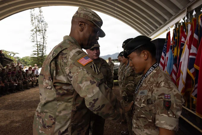 A military officer is awarding a medal to another uniformed individual during a formal ceremony. Multiple flag drapes are visible in the background, and a group of people in uniform watch the event. The ceremony takes place under an arched structure surrounded by trees.