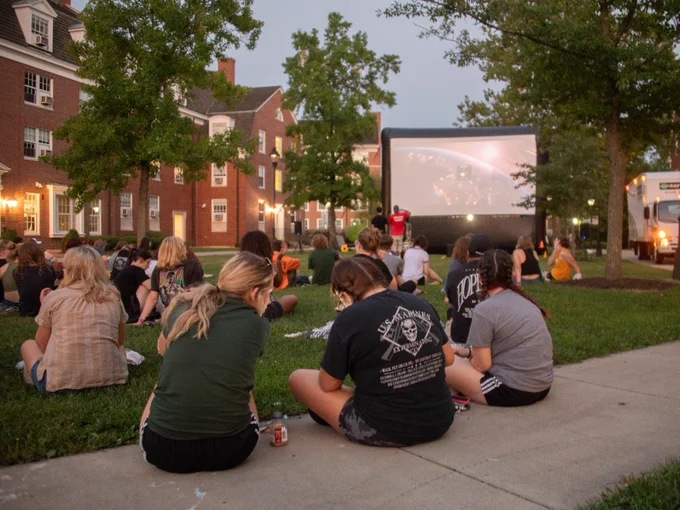 Students sit on the West Green lawn at Ohio University watching a movie on a large screen. 