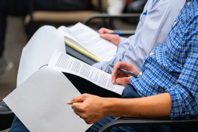 People seated closely, reviewing printed documents and taking notes during a meeting or seminar.