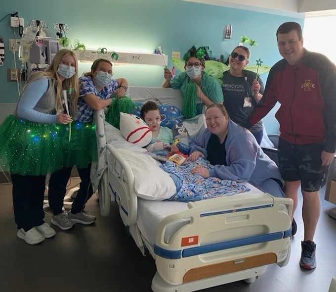 A hospital room with a patient in bed surrounded by five individuals. The patient is holding a 'Cat in the Hat' book. The group is dressed in festive attire, with green clothing and accessories. One person is wearing an Iowa State jacket. Smiles are shared by all.