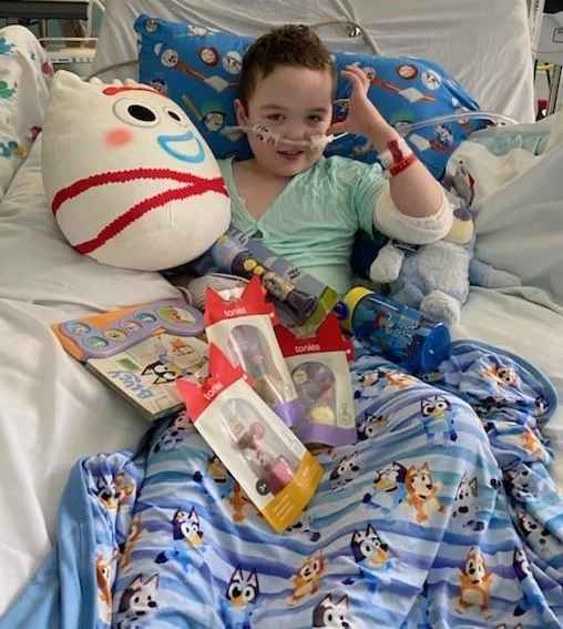 A person is sitting in a hospital bed surrounded by "Bluey" merchandise, including a blanket and toys. A large "Frozen" Olaf plush is also on the bed. The person has nasal tubes and appears to be enjoying the toys.