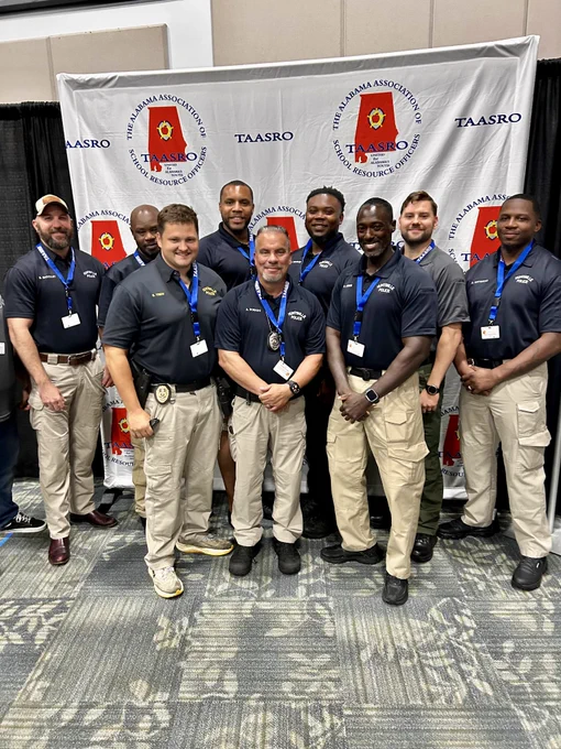 A group of individuals wearing dark polo shirts with badges and khaki or dark pants is standing together in front of a backdrop displaying the logo of the Alabama Association of School Resource Officers (TAASRO). The backdrop features repeated text and graphics of the organization’s emblem. The group is posed for a formal photo at what appears to be an event or conference.