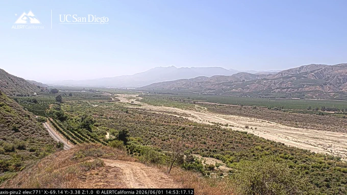 A dry scrubby valley landscape with a dirt road is in the forground, a dry stream bed is in the middle ground, and distant agricultural fields at the foot of mountains. The sky is blue with no clouds. ALERTCalifornia and UC San Diego's logos are in the upper left corner. 