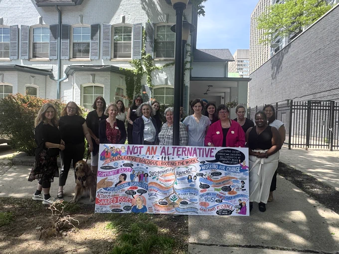 A group of people stand outside a building on a sunny day, smiling together behind a large, colorful graphic poster that reads "NOT AN 'ALTERNATIVE'".