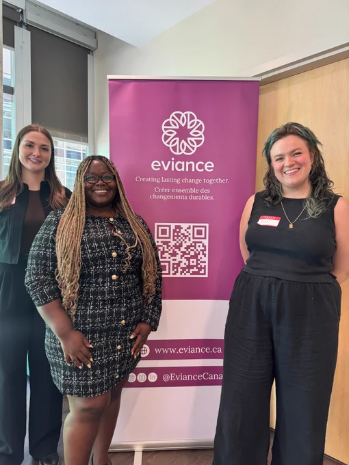 Three women stand smiling next to a purple roll-up banner that reads "eviance: Creating lasting change together."
