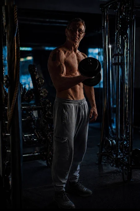 Standing in a dark gym holding a dumbbell after training, dramatic lighting highlighting muscle definition and a quiet, focused atmosphere.