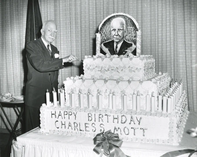 C.S. Mott cuts into a large birthday cake that says "Happy birthday Charles S. Mott" and features a portrait of himself at the top.