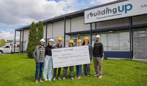 A group of seven people wearing hard hats stand smiling in front of a building labeled "Building Up." They are holding a large check, indicating a donation or partnership. The mood is cheerful and collaborative.