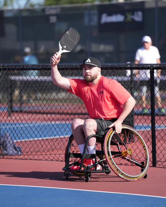 A man in a wheelchair playing pickleball, holding a paddle up, wearing a red shirt and cap on an outdoor court.