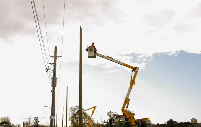 Toronto Hydro workers use a bucket truck to install or repair a wooden hydro pole with overhead power lines under a cloudy sky.