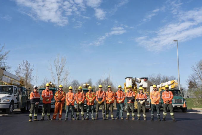 Toronto Hydro crew members stand in a line wearing high-visibility safety gear and hard hats, with utility bucket trucks and equipment parked behind them under a clear blue sky.