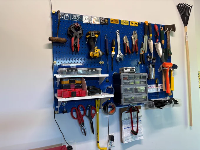 Blue Wall Control metal pegboard mounted on a garage wall, neatly organizing a variety of tools and accessories. The setup includes a cordless drill, wrenches, pliers, clamps, trowels, an axe, hammers, scissors, storage bins, shelves, and small parts organizers. A rake hangs beside the pegboard, while hooks, shelves, and accessory holders maximize vertical storage and keep the workspace clean, organized, and easily accessible. Photo courtesy - Joe.