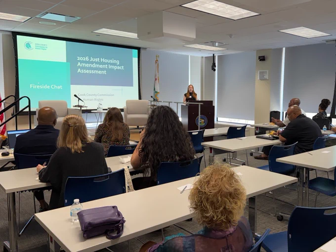 Claire Dillon, an individual with shoulder length red hair stands behind a podium. Claire is presenting on the findings of the Just Housing Amendment Impact Assessment.