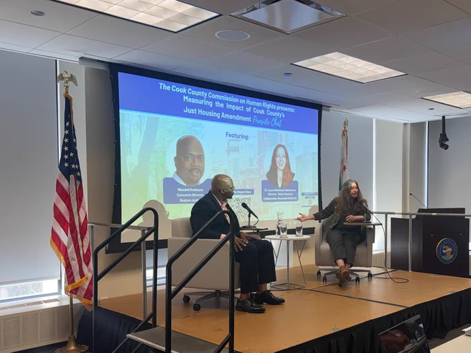 
Wendell Robinson sits on stage across from Dr. Laura Nussbaum-Barberena. The background image includes the promotional flyer. The American flag and Cook County flag stand behind the stage. 
