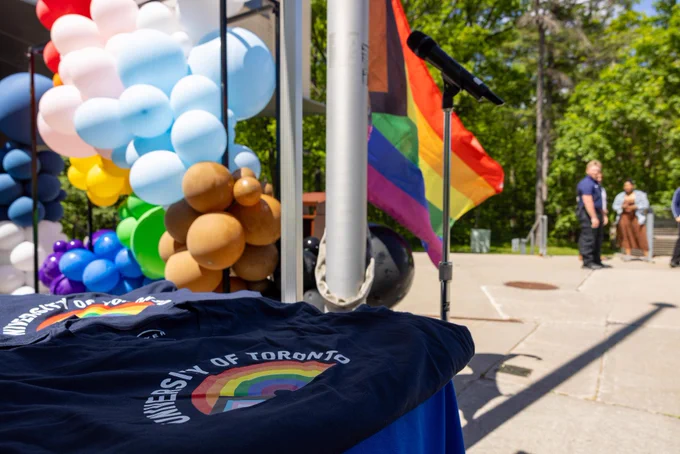 A table with a "University of Toronto" T-shirt featuring a rainbow design is in the foreground. A microphone stand and a large rainbow flag are nearby. Colourful balloons in various shapes and colours, including white, blue, and brown, decorate the area. People are in the background, with greenery visible.