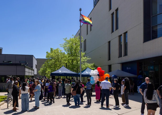 A group of people gather outside a modern building with large windows. Several tents are set up, adorned with balloons. A flagpole displays the Progress Pride flag. The scene is set on a clear, sunny day.