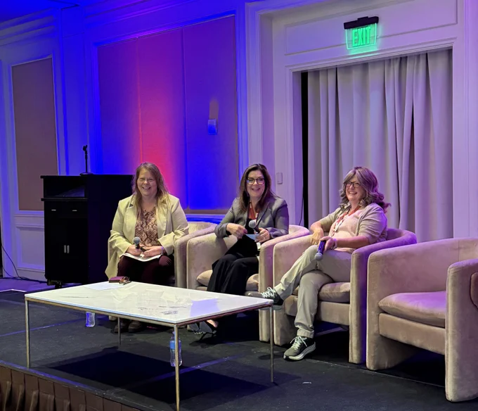 A photo of three businesswomen sitting in three chairs on a stage with humored facial expressions. There is a table in front of them with a red wallet/purse on it. Behind them is a wall with blue and red ambient lighting. Above the curtains in the door way is a green exit sign.