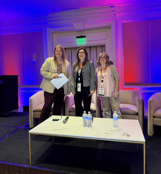 1. Photo of three businesswomen standing together on a stage. There is a table in front of them with three water bottles and three microphones. Behind them are a set of four chairs. The wall behind them is lit up with blue and red ambient lighting and there is an LED exit sign above the curtains in the doorway.