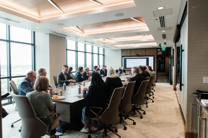 A group of people are seated around a long conference table in a modern meeting room with large windows. A presentation is displayed on a screen at the far end of the room.
