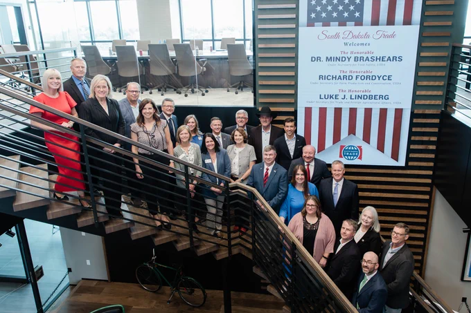 A group of people dressed in formal attire stand on a staircase in a modern building. In the background, a large banner welcomes Dr. Mindy Brashears, Richard Fordyce, and Luke J. Lindberg. An American flag is featured on the banner.