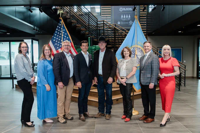 Eight people are standing in an office lobby in front of the United States and South Dakota flags. They are dressed in business and casual attire. A staircase and a reception area are visible in the background.